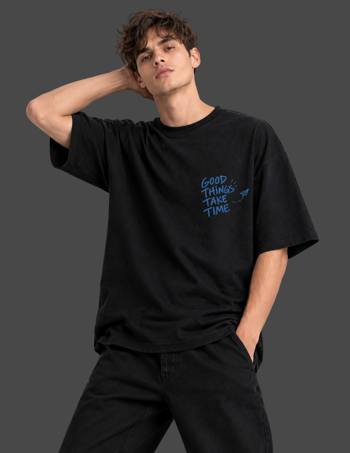 A man wearing a black t-shirt with a large graphic print on the back, sitting on a white block against a dark gray background. The graphic features a blue square with the text 'GOOD THINGS TAKE TIME' and small illustrations of people.
