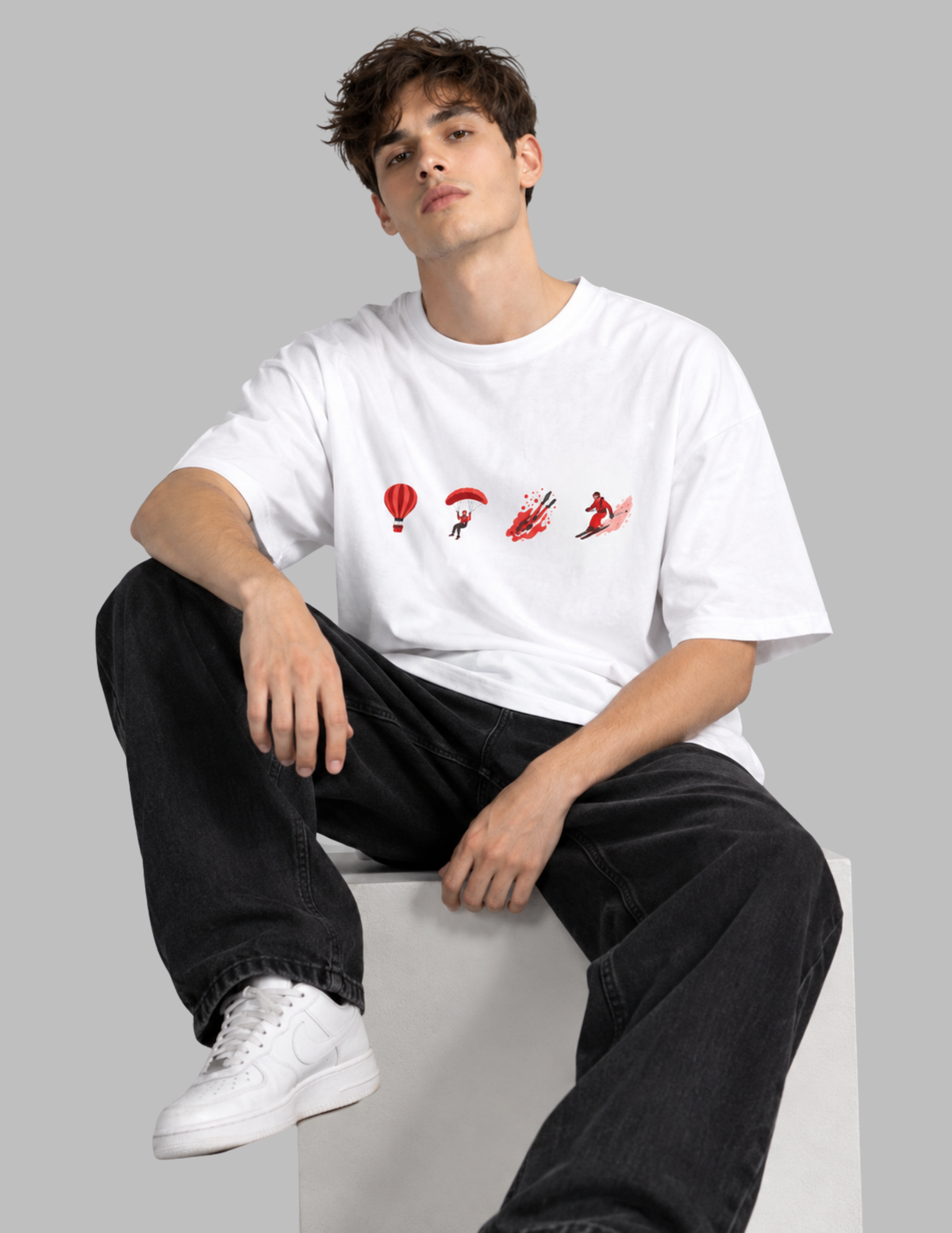 A young man sitting on a white block wearing a white t-shirt with a red graphic print of parachutists, paired with black jeans and white sneakers, against a solid gray background.