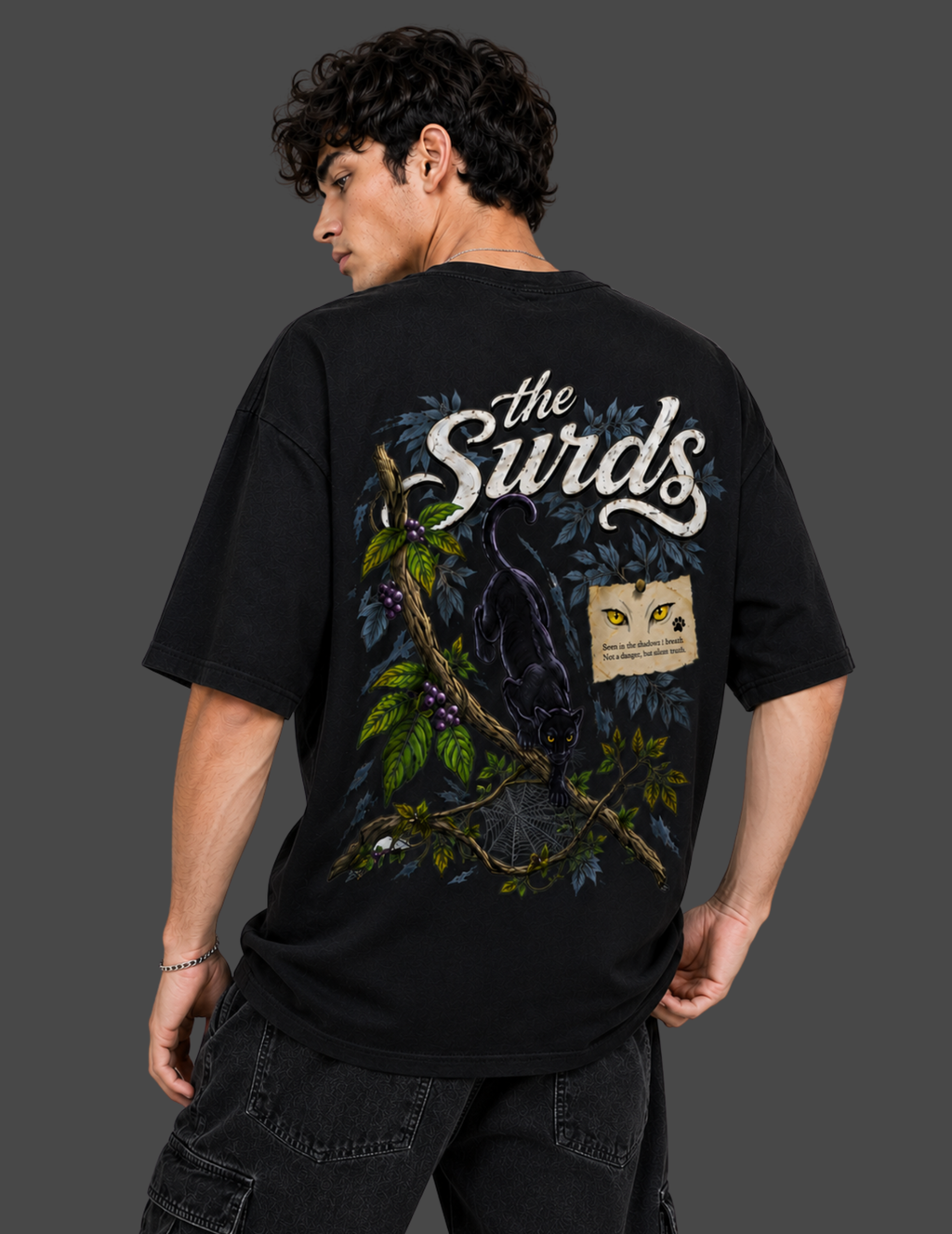 A man wearing a black graphic t-shirt from the back, featuring a large design with the text 'the Surds' and an illustration of a black cat on a branch with green leaves and a small yellow-and-white patch graphic.