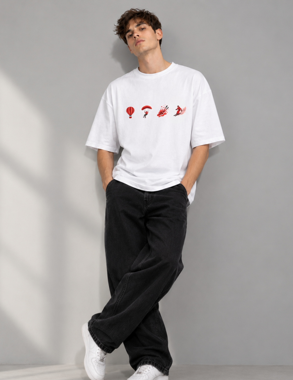 A man wearing a white crewneck t-shirt with a red graphic print of four small figures across the chest, paired with loose black jeans and white sneakers, standing against a gray wall.