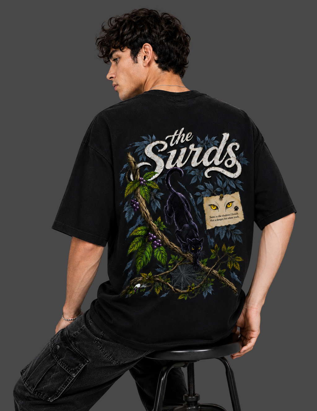 A man wearing a black graphic t-shirt with a large back print featuring the text 'the Surds' and an illustration of a black cat under a tree, seated on a stool against a dark gray background.