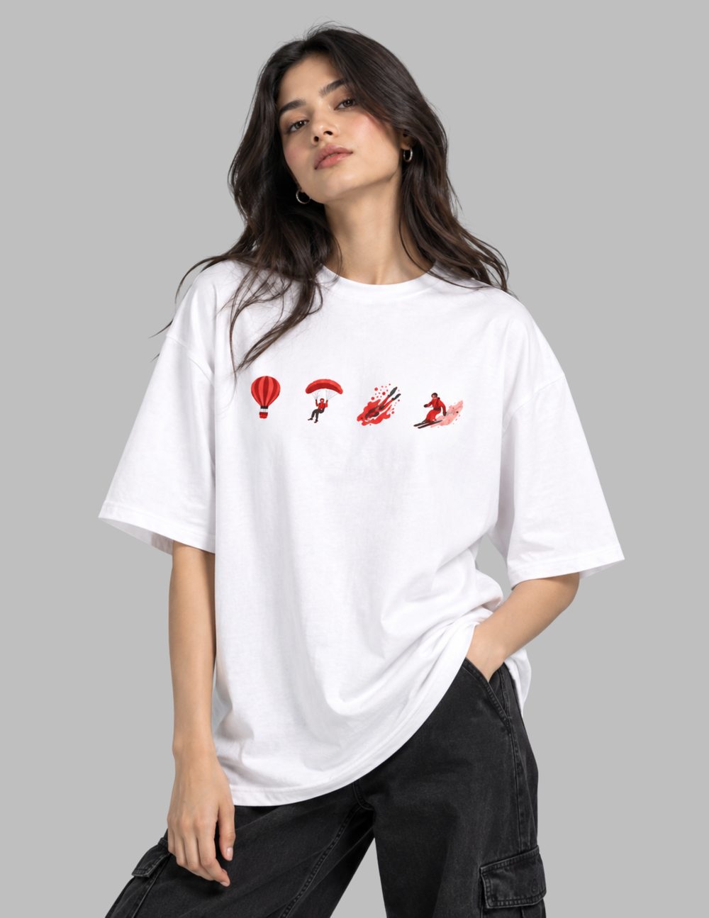 A woman wearing an oversized white t-shirt with a red graphic print of parachutist silhouettes, paired with black cargo pants, against a solid gray background.