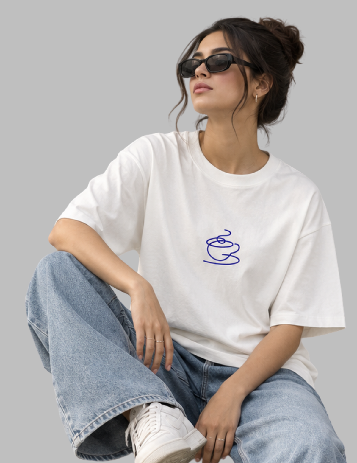 A woman wearing a white oversized t-shirt with a blue coffee cup outline graphic on the chest, paired with light-wash blue jeans and white sneakers, posed against a solid gray background.