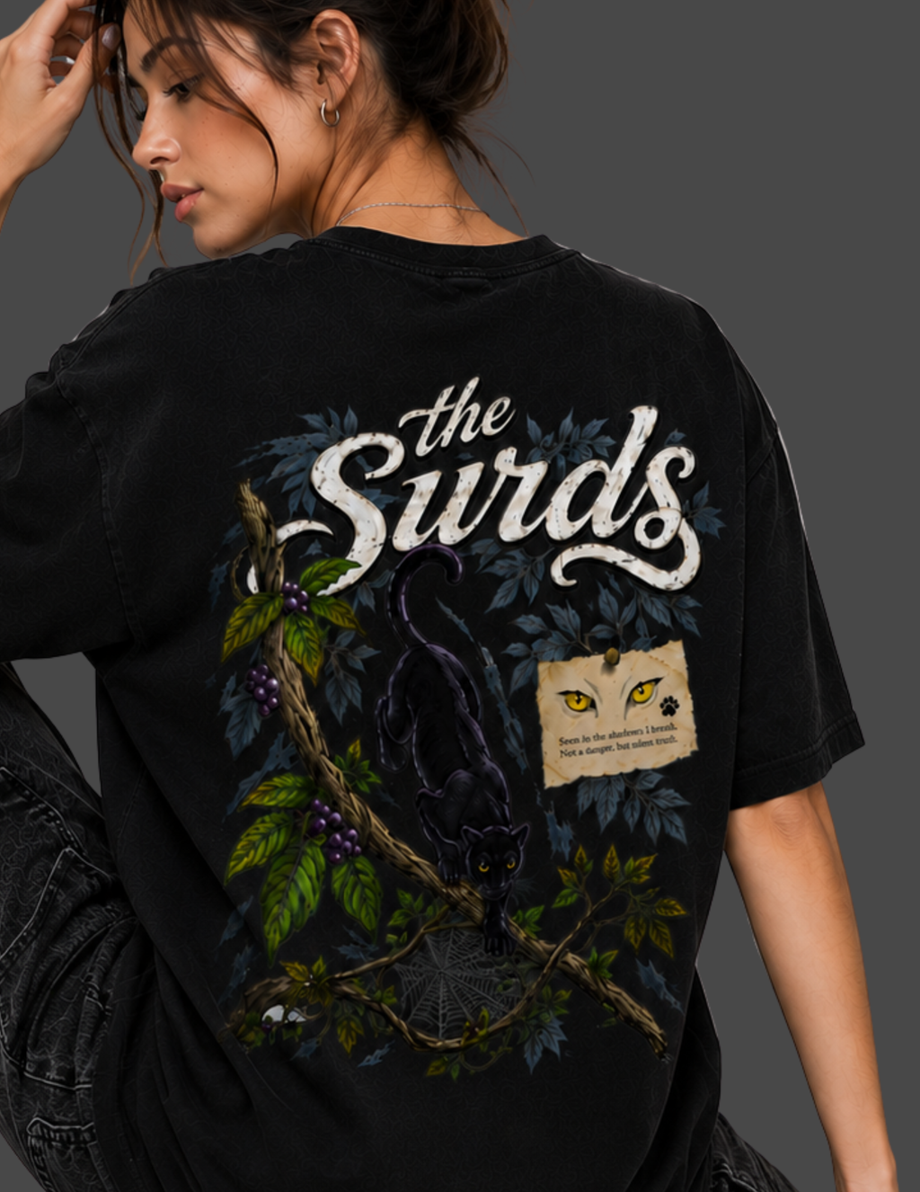 A woman wearing a black graphic t-shirt with a large back print featuring the text 'the Surds', a black cat on a branch, green leaves, and a small yellow-eyed card graphic, shown against a dark gray background.