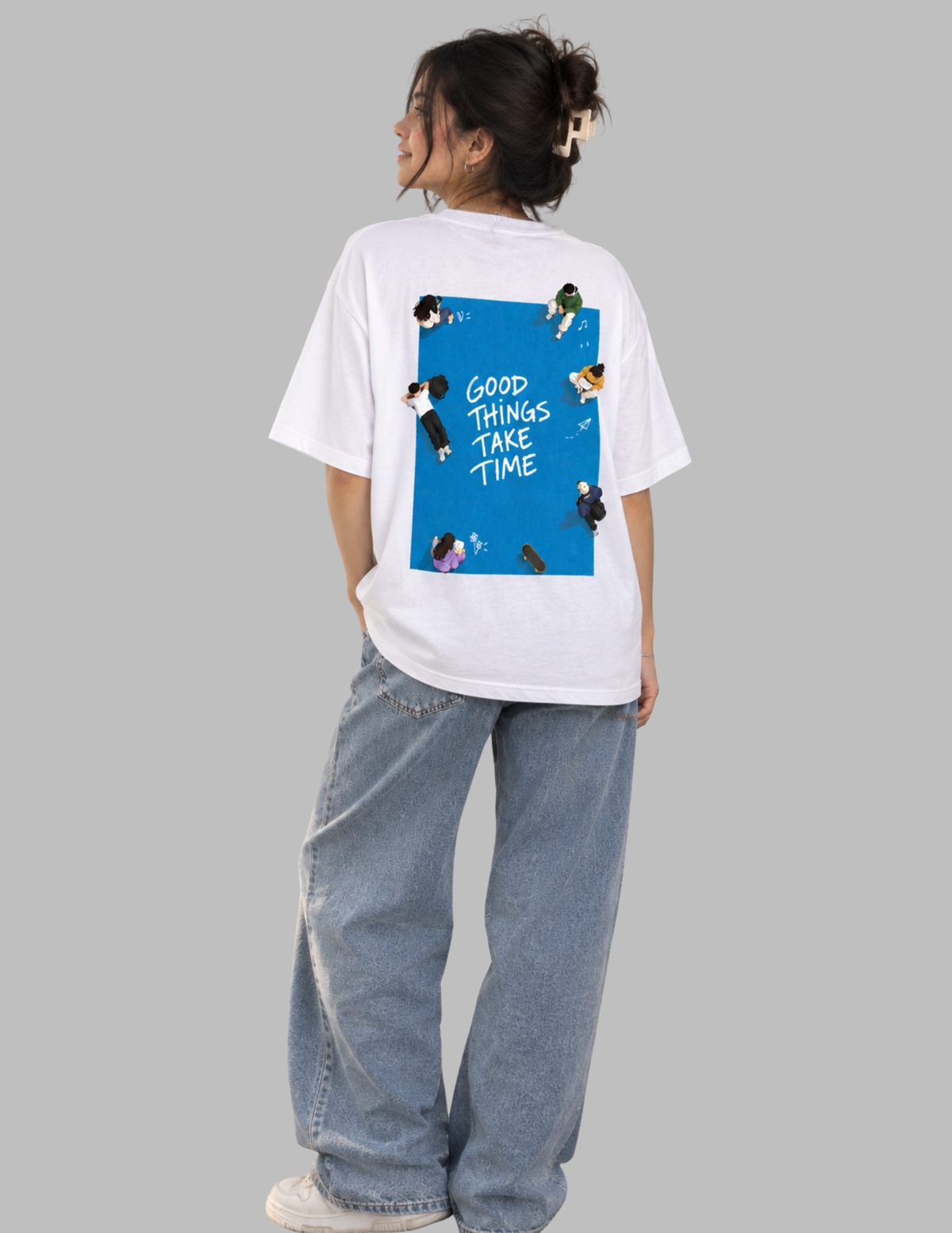 A model wearing a white oversized t-shirt with a blue graphic print on the back, paired with light-wash wide-leg jeans, shown from a rear three-quarter view against a solid gray background.