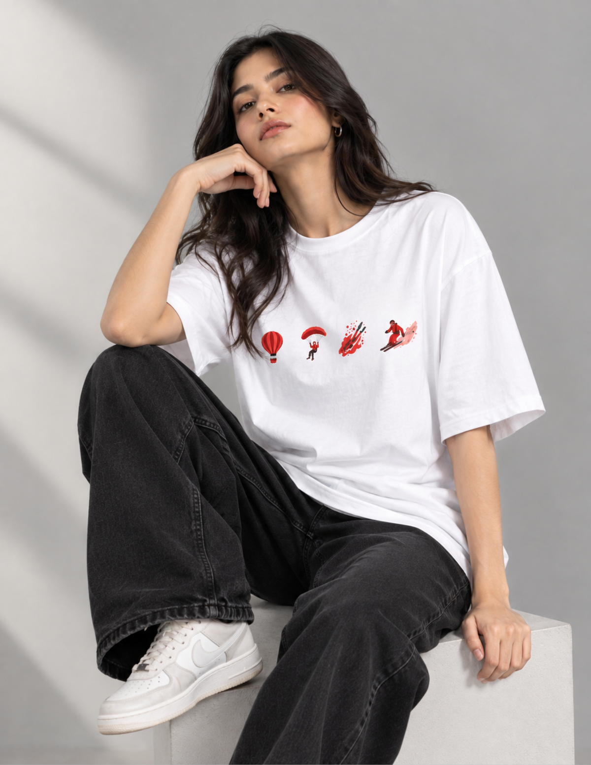 A woman sits on a white block wearing a white graphic t-shirt with red and black skydiving-themed illustrations, black wide-leg jeans, and white sneakers against a gray studio background.