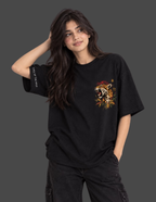 A woman modeling a black oversized t-shirt with a colorful tiger graphic on the left chest and white text on the right sleeve, paired with black cargo pants against a dark gray background.