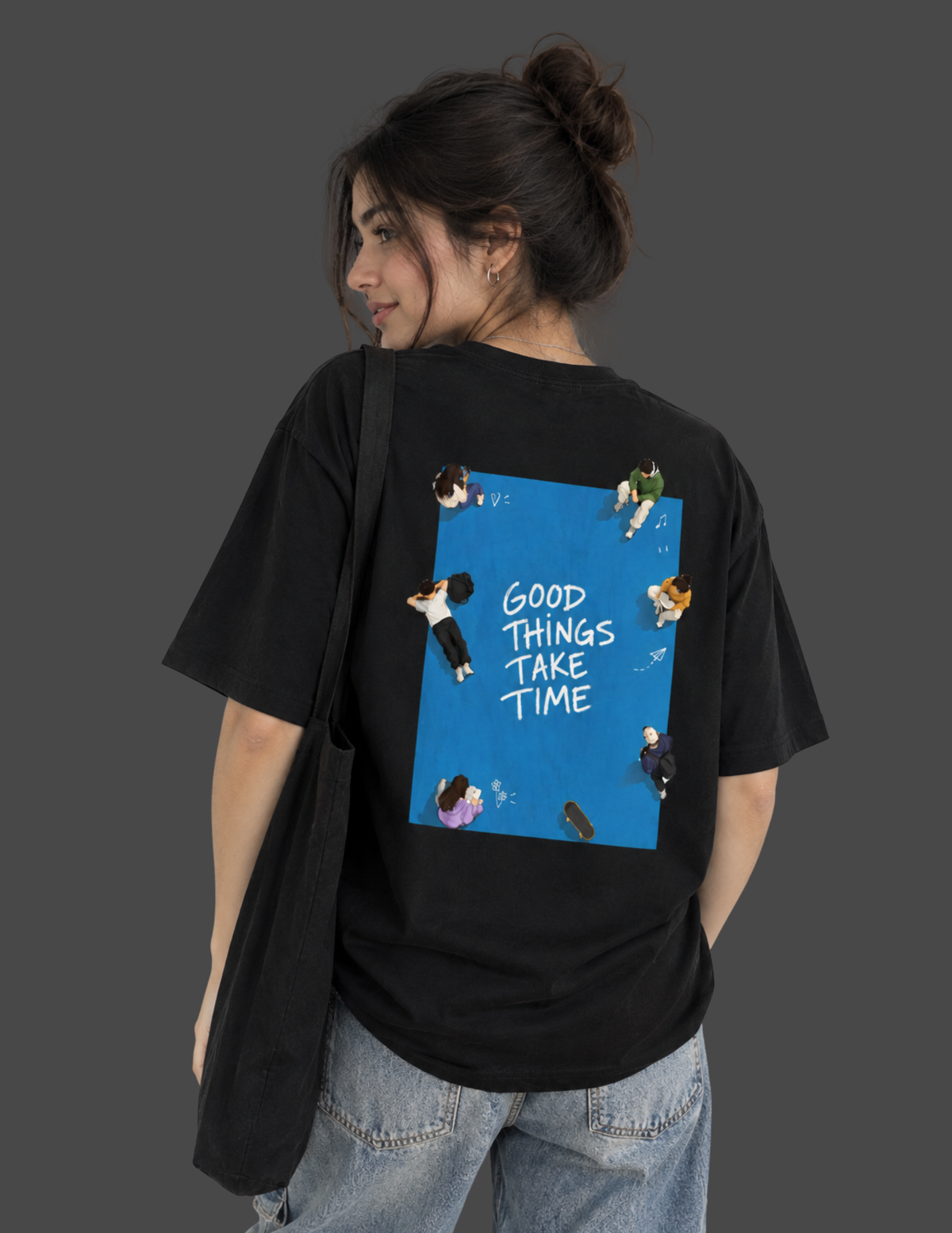 A woman wearing a black oversized t-shirt from the back, featuring a blue graphic print with illustrations of people and the text 'GOOD THINGS TAKE TIME'. She is also wearing light-wash blue jeans and carrying a black shoulder bag against a solid dark gray background.