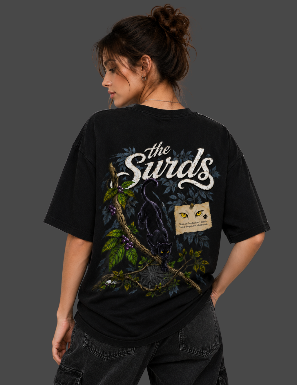 A woman shown from the back wearing a black graphic t-shirt with 'the Surds' printed in white script, featuring a dark bird, green leaves, and a small yellow-and-black patch graphic.