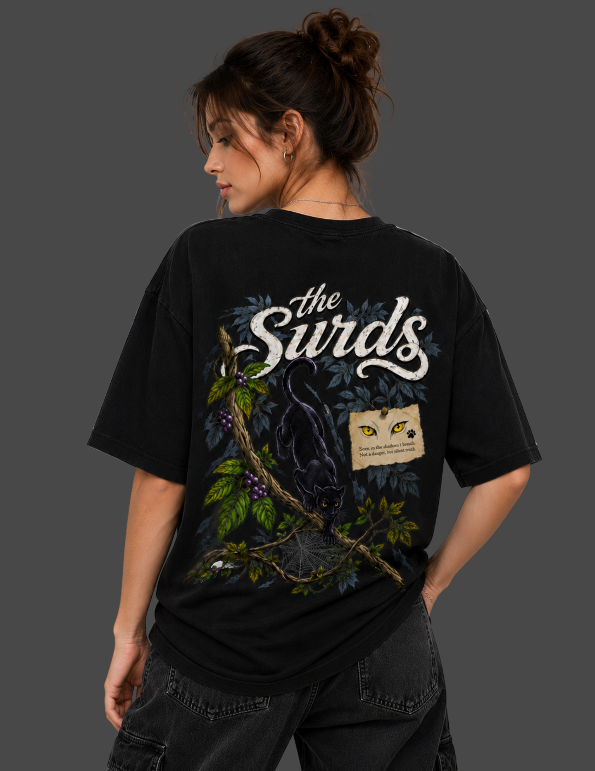 A woman shown from the back wearing a black graphic t-shirt with 'the Surds' printed in white script, featuring a dark bird, green leaves, and a small yellow-and-black patch graphic.