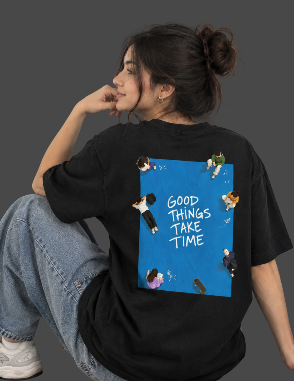 A woman wearing a black oversized t-shirt with a large graphic print on the back, sitting against a dark gray background. The graphic features a bright blue square with the text 'GOOD THINGS TAKE TIME' and small illustrated figures.