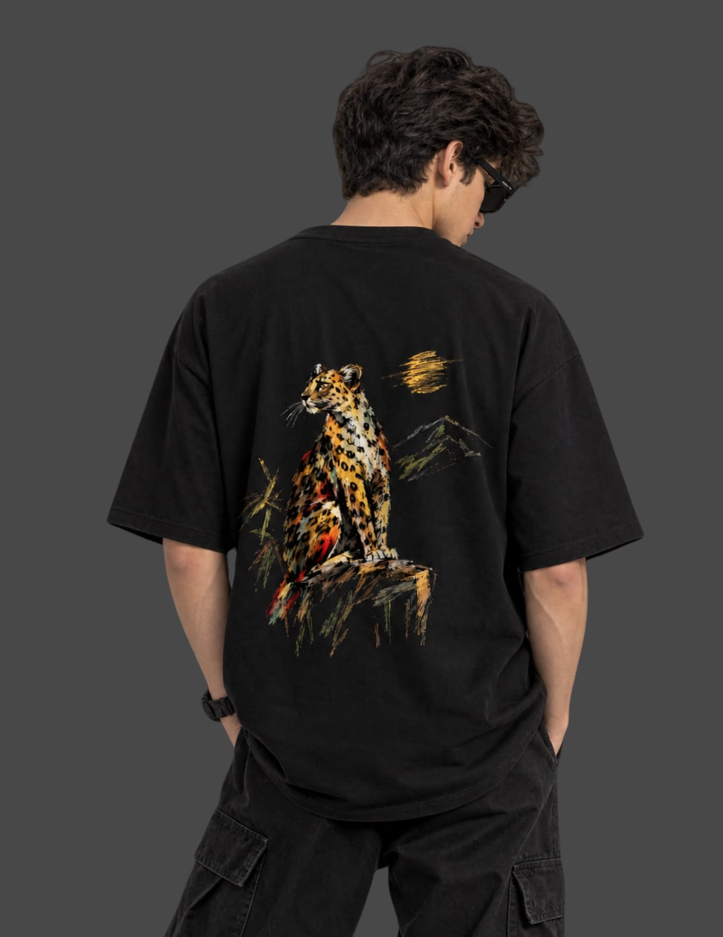 A man viewed from behind wearing a black t-shirt with a large graphic print of a leopard sitting on a rock with a sunset behind it, set against a dark gray background.