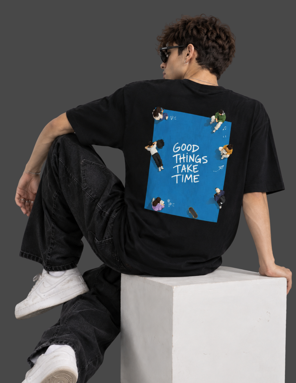 A man wearing a black t-shirt with a large graphic print on the back, sitting on a white block against a dark gray background. The graphic features a blue square with the text 'GOOD THINGS TAKE TIME' and small illustrations of people.