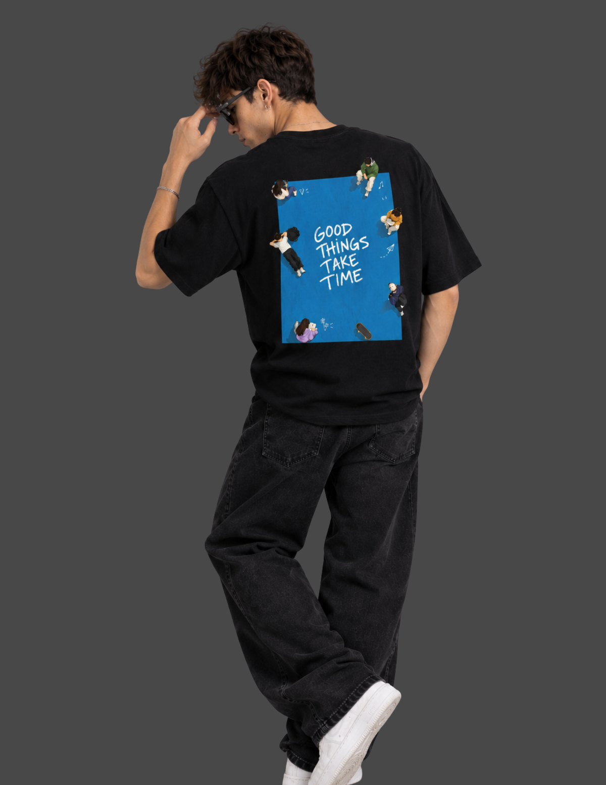 A man wearing a black t-shirt with a large graphic print on the back, sitting on a white block against a dark gray background. The graphic features a blue square with the text 'GOOD THINGS TAKE TIME' and small illustrations of people.