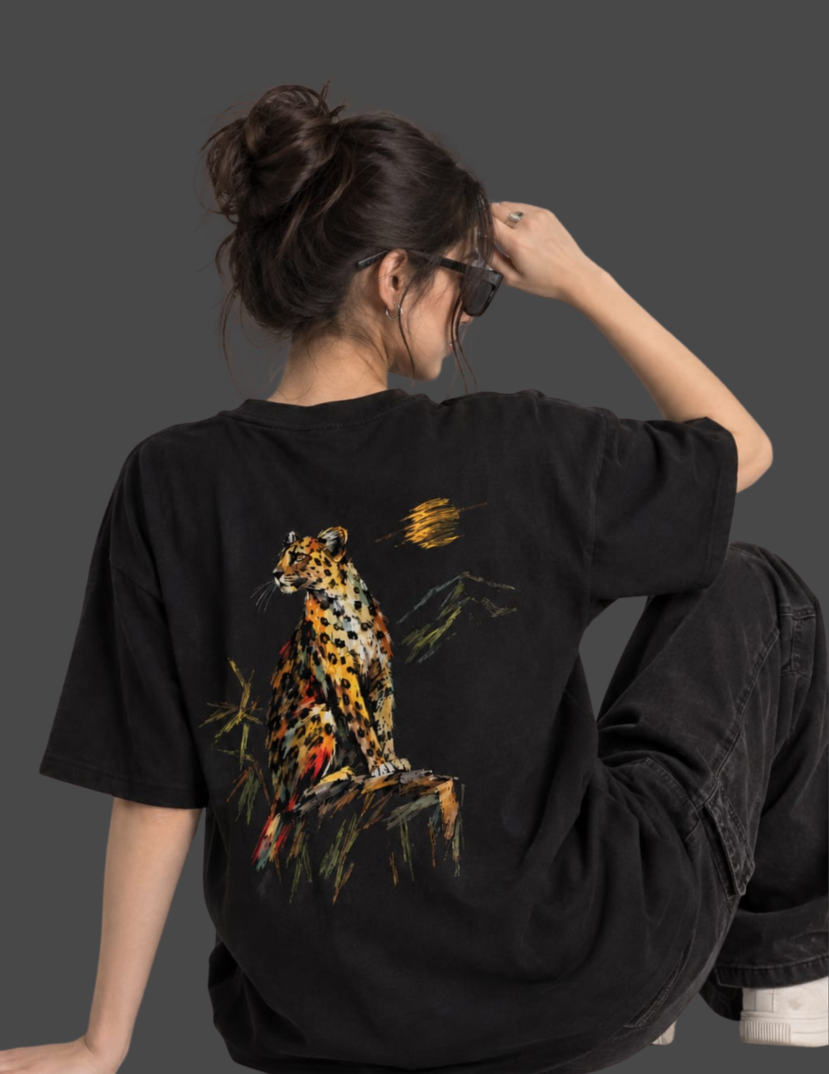 A woman shown from behind wearing a black oversized graphic T-shirt with a large leopard print on the back, paired with dark jeans, against a dark gray studio background.