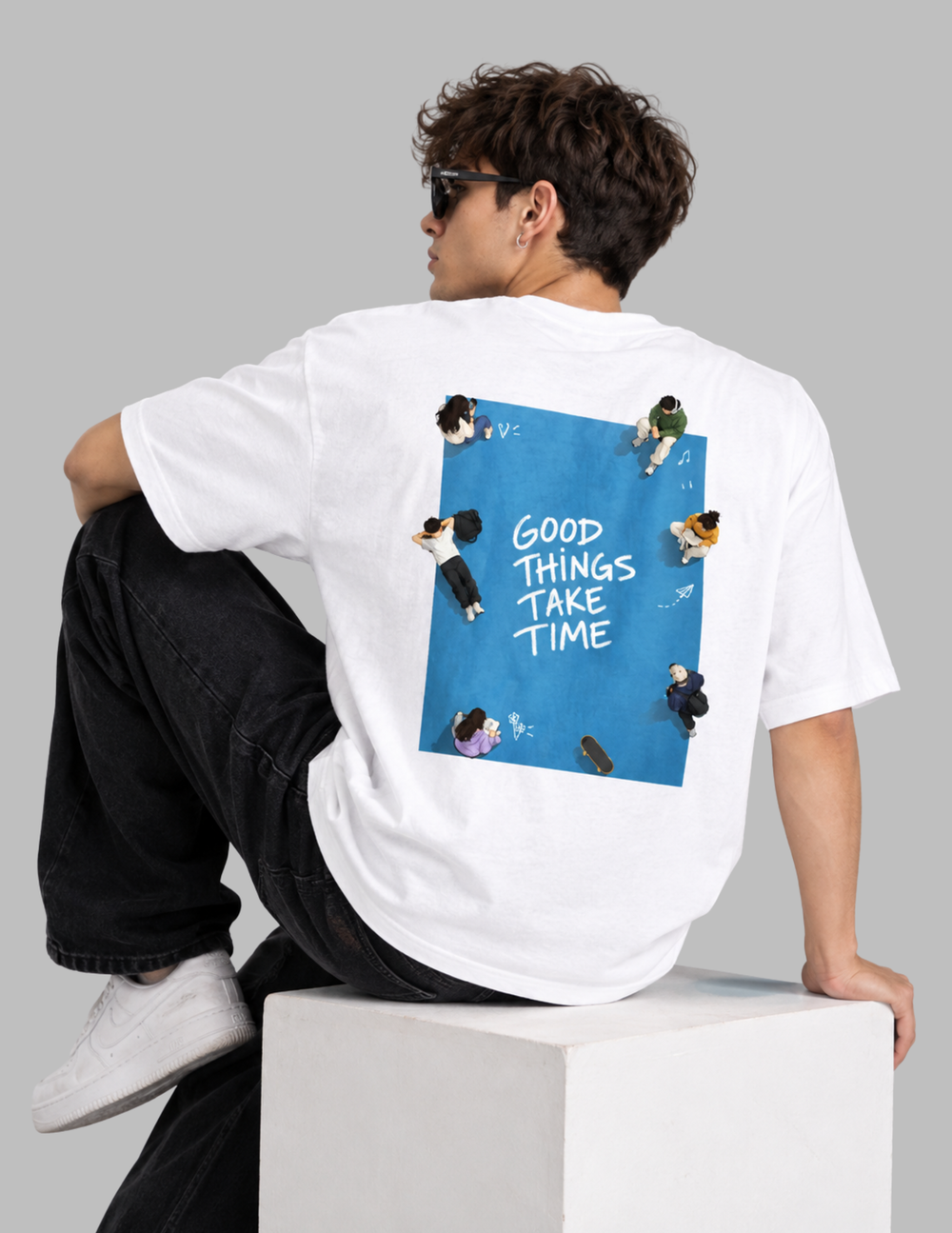 A man wearing a black t-shirt with a large graphic print on the back, sitting on a white block against a dark gray background. The graphic features a blue square with the text 'GOOD THINGS TAKE TIME' and small illustrations of people.