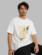 A man wearing a white short-sleeve t-shirt with a vintage-style graphic print on the front featuring a peacock feather and a book illustration, paired with black pants, set against a solid gray background.