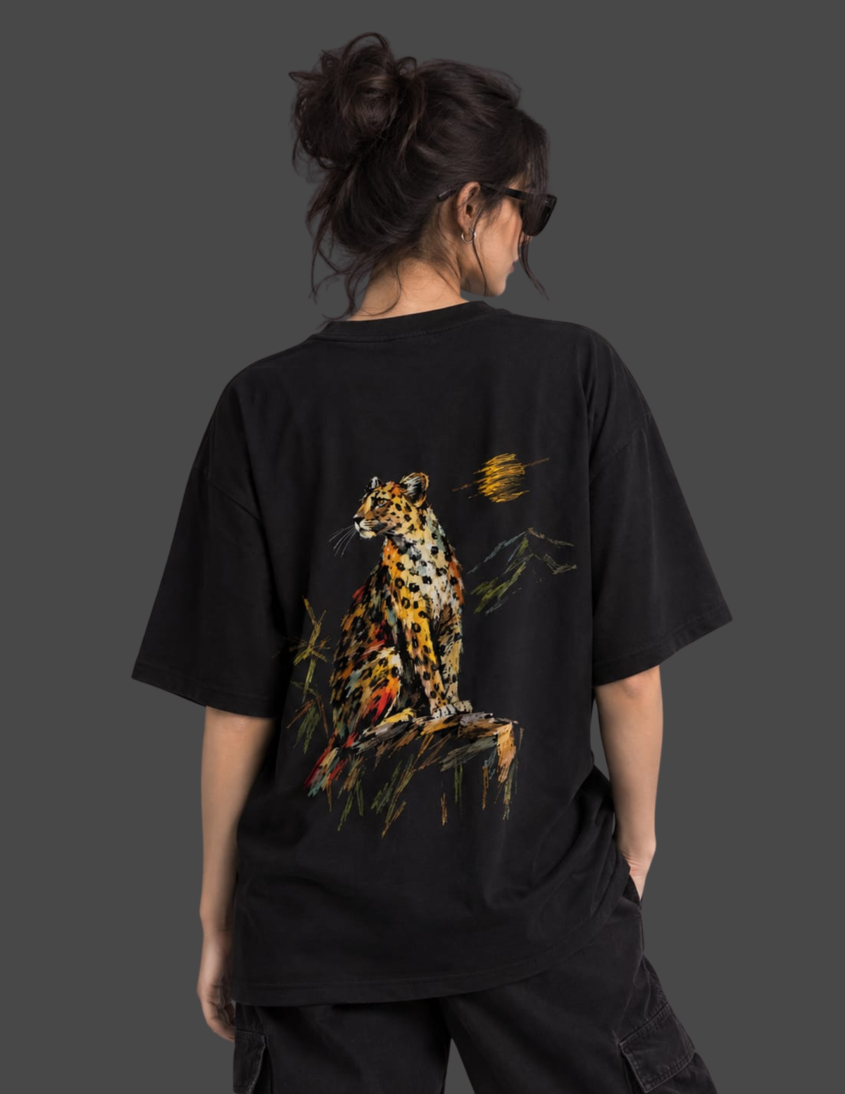 A person shown from the back wearing an oversized black T-shirt with a large colorful leopard graphic on the back, photographed against a dark gray studio background.