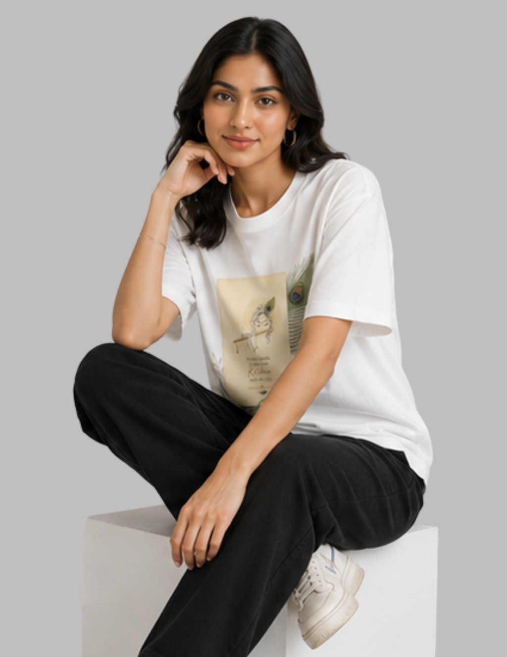 A woman sits on a white block wearing a white graphic t-shirt, black pants, and white sneakers against a solid gray background.