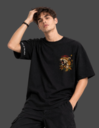 A man wearing a black oversized t-shirt with a colorful leopard graphic on the left chest and white text on the right sleeve, posed against a dark gray studio background.