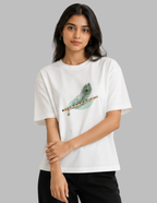 A woman modeling a white short-sleeve crewneck t-shirt featuring a graphic of a teal and green peacock feather resting on a brown flute. She is wearing black pants against a solid gray studio background.