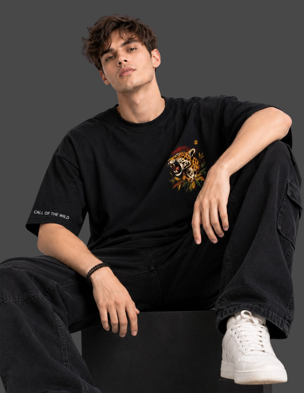 A man wearing a black graphic t-shirt with a tiger print on the chest and 'CALL OF THE WILD' text on the sleeve, paired with black cargo pants and white sneakers, sitting against a dark gray background.