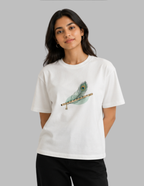 A woman modeling a white short-sleeve crewneck t-shirt featuring a graphic of a teal and gold peacock feather resting on a wooden flute. She is wearing black pants against a solid gray studio background.