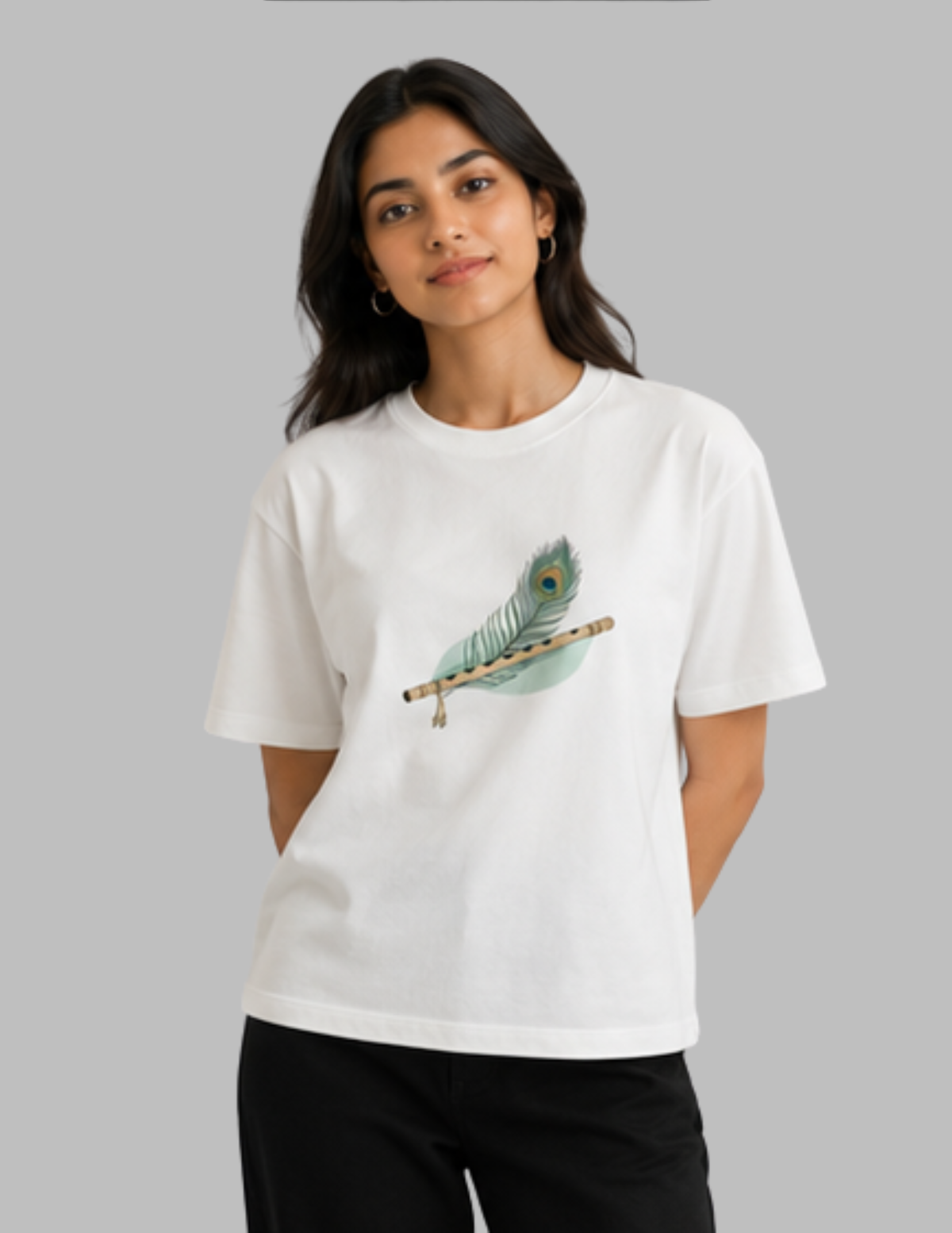A woman modeling a white short-sleeve crewneck t-shirt featuring a graphic of a teal and gold peacock feather resting on a wooden flute. She is wearing black pants against a solid gray studio background.