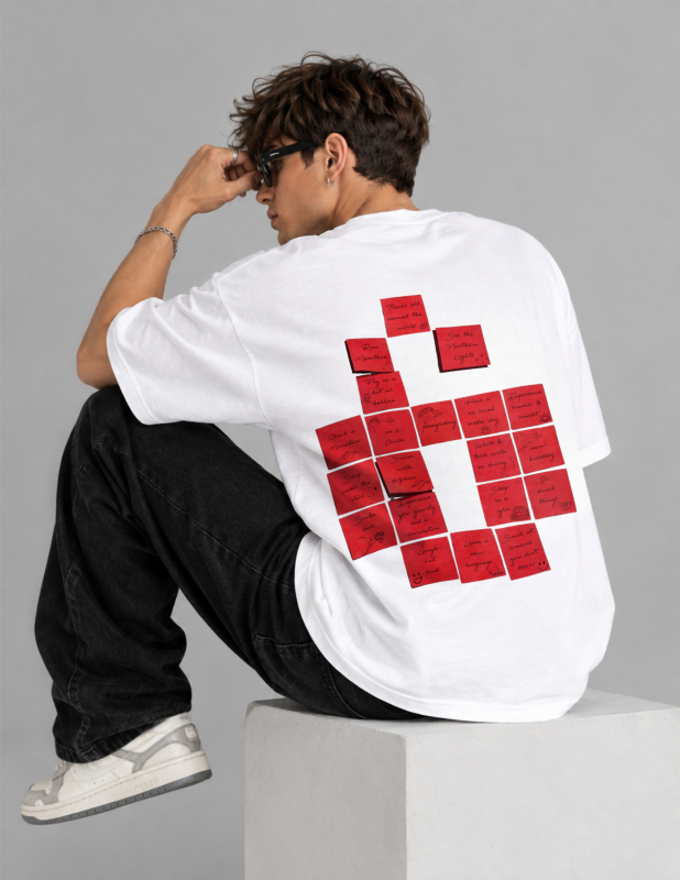 A man wearing a white oversized T-shirt with a large red graphic made of rectangular sticky-note-like shapes on the back, seated on a white block against a light gray studio background.