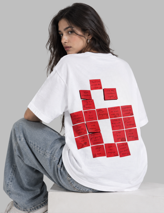 A woman wearing a white oversized T-shirt with a large red graphic made of rectangular sticky-note-like shapes on the back, paired with light blue jeans, seated against a light gray background.