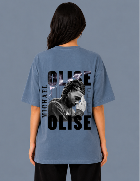 Person wearing a blue t-shirt with 'OLISE' and 'MICHAEL' printed on the back against a gray background