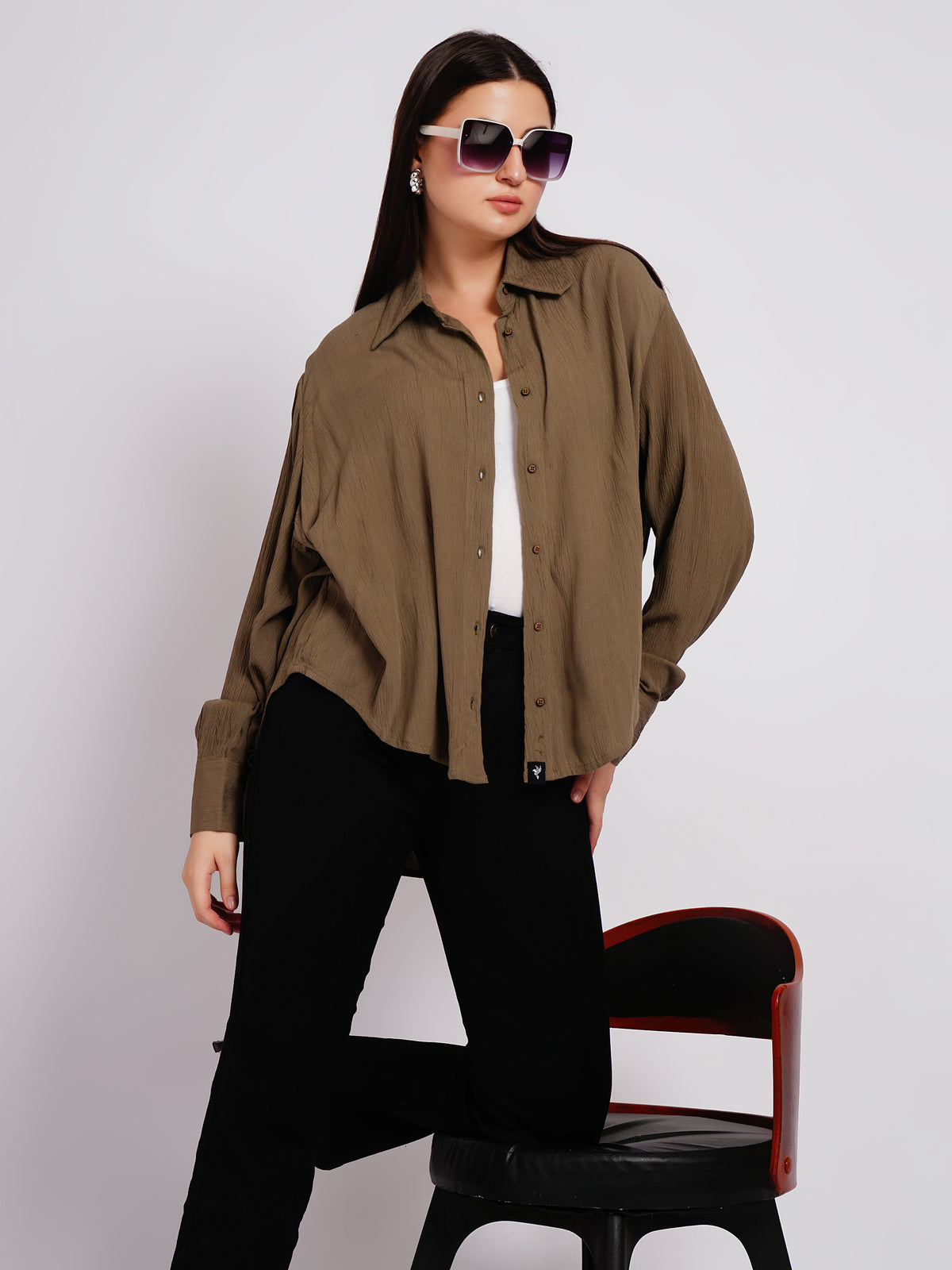 Woman wearing an olive green shirt and black pants, standing next to a chair against a plain background.