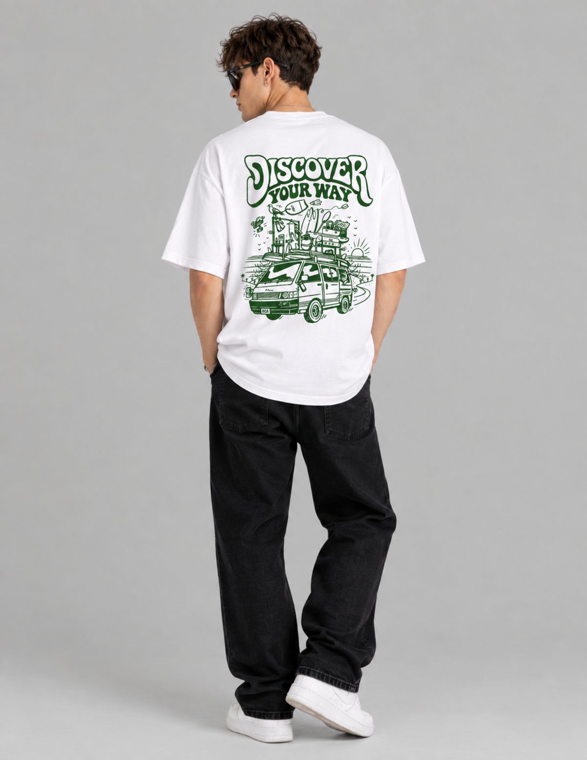 A man viewed from behind wearing a white t-shirt with a green graphic print that reads 'DISCOVER YOUR WAY' above an illustration of a car, paired with black jeans and white sneakers, against a solid gray studio background.