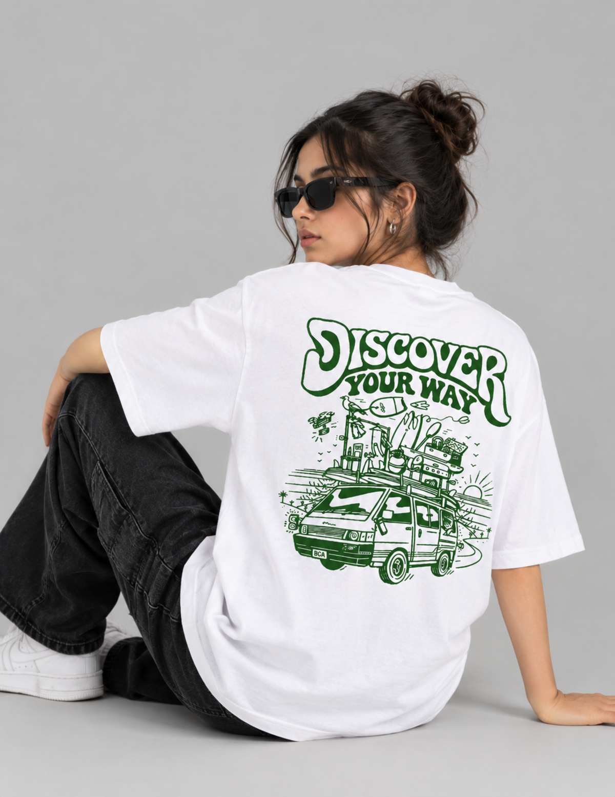 A woman wearing a white oversized T-shirt with a green graphic print on the back, paired with black jeans and white sneakers, seated against a light gray studio background.