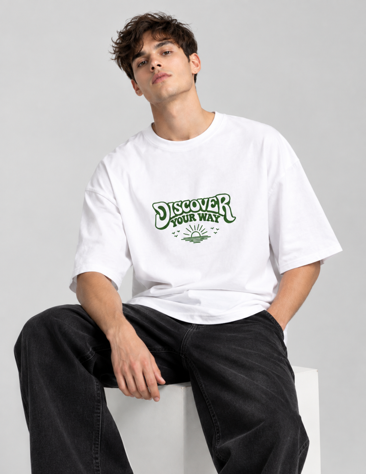 A man wearing a white oversized t-shirt with a green 'Discover Your Way' graphic on the chest, paired with black jeans, sitting on a white block against a light gray background.
