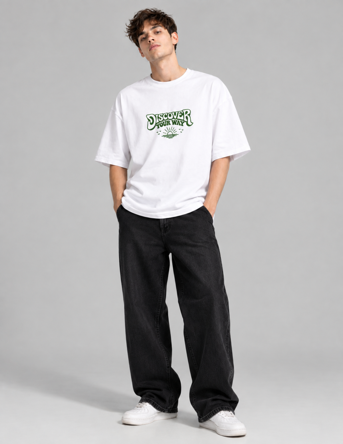 A man modeling a white crewneck t-shirt with a green 'Discover Gamer' graphic on the chest, paired with black jeans and white sneakers, against a solid gray studio background.