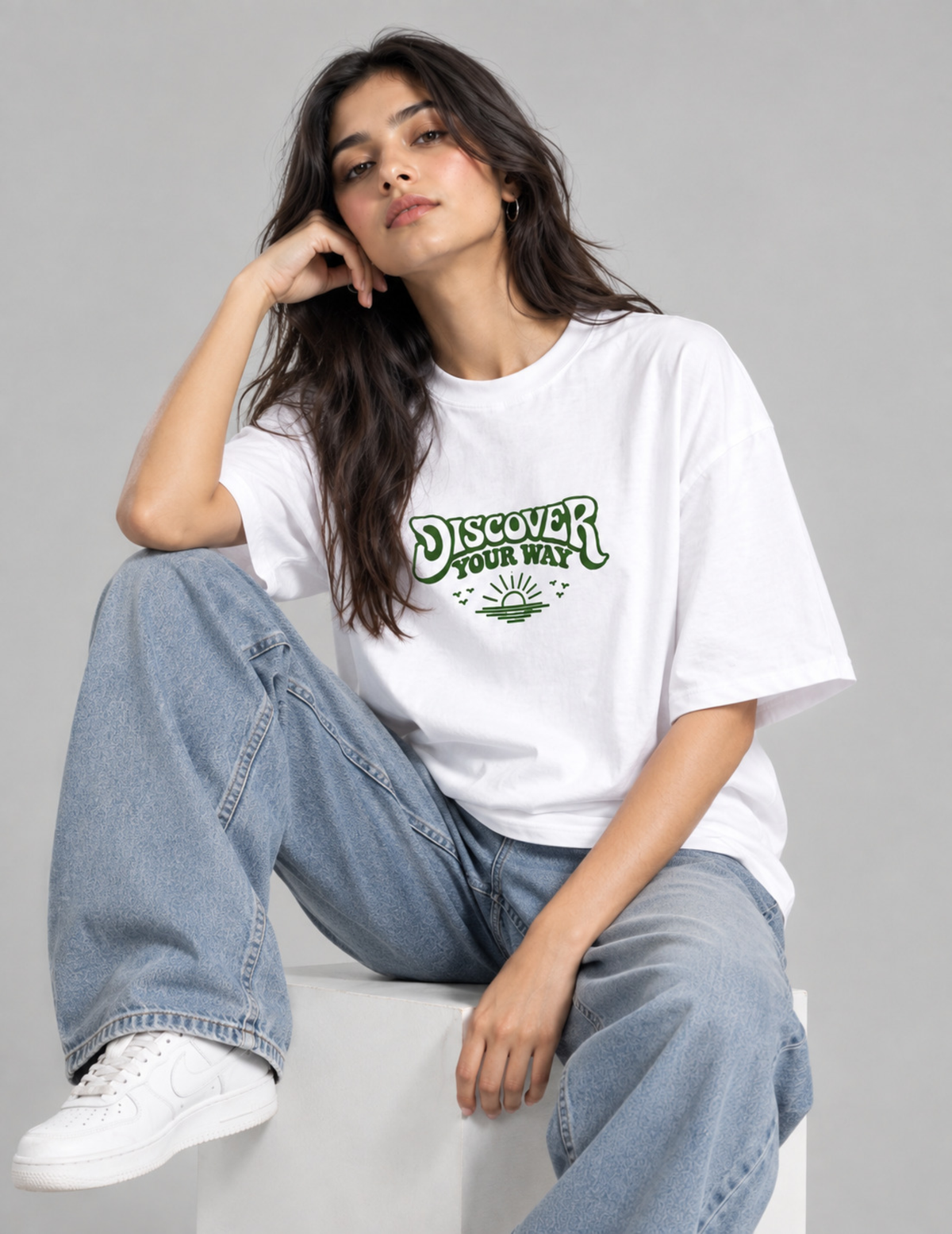 A woman wearing a white graphic t-shirt with green text that reads 'DISCOVER YOUR WAY' and light blue jeans, sitting on a white block against a gray studio background.