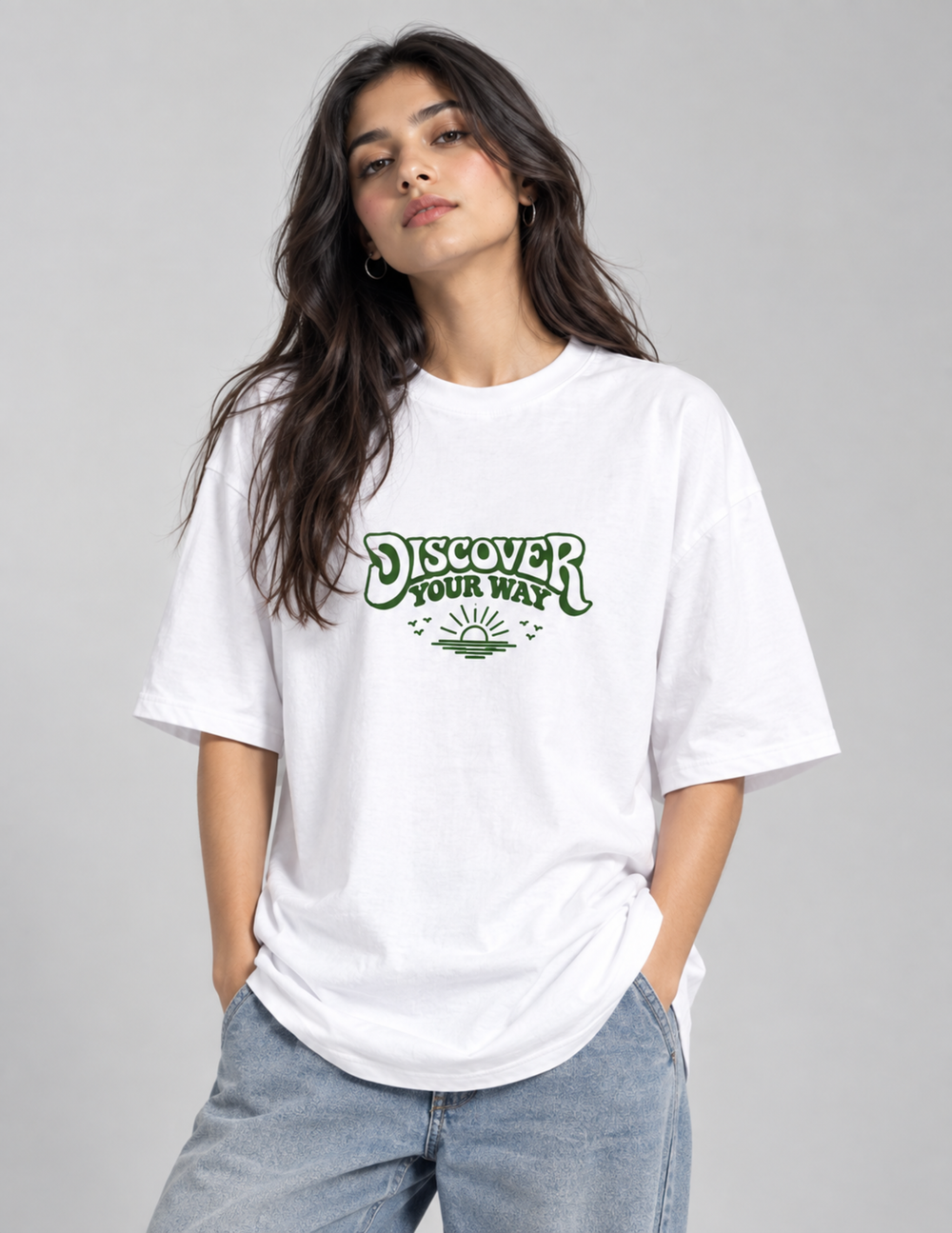 A woman wearing a white oversized t-shirt with a green retro-style graphic that reads 'DISCOVER YOUR WAY' and a sun design, paired with light-wash jeans, against a solid gray studio background.