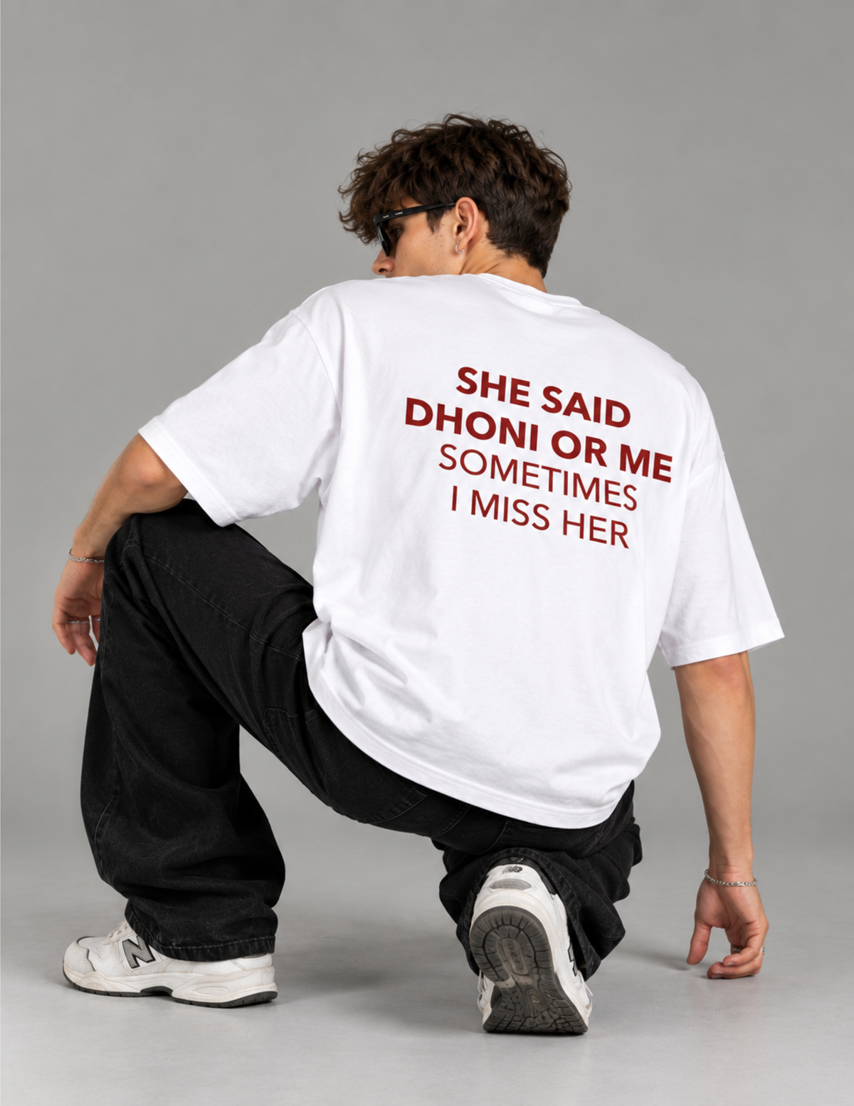 A man crouching from behind wearing a white oversized t-shirt with red text that reads 'SHE SAID DHONI OR ME SOMETIMES I MISS HER' on the back, paired with black pants and white sneakers, against a solid gray studio background.