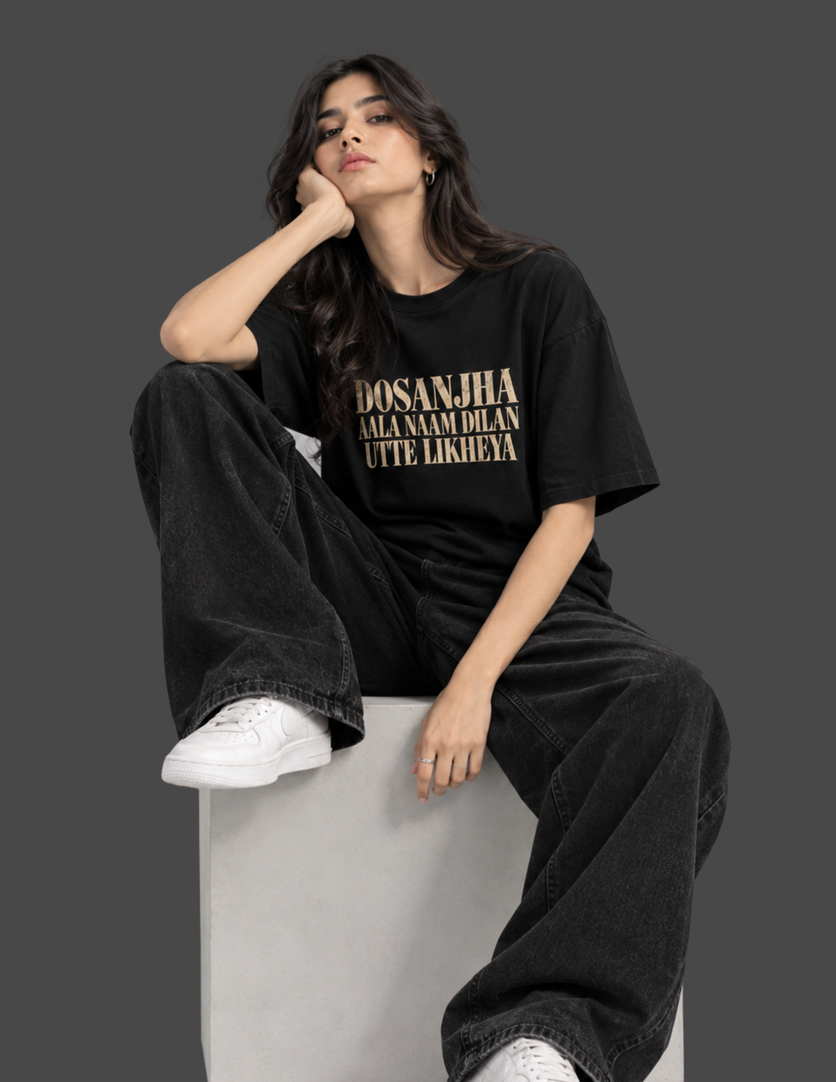 A woman wearing a black oversized t-shirt with a large graphic print on the back, sitting on a concrete block against a dark gray studio background.